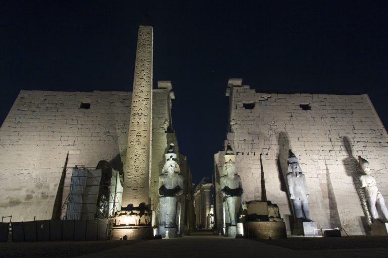 Image of Luxor Temple at night. The columns and Obelisk are lit up. The sky in the background is dark.