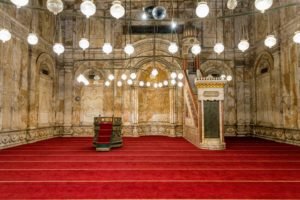 Inside of the Mosque of Mohammed Ali, Red carpet floors and globe lights that are on hanging form the ceiling. The Pulpit is in the background.