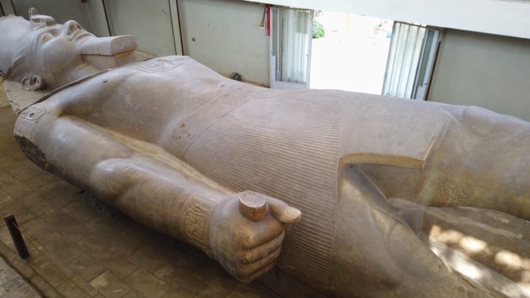 Photo of the large Ramessess the Second statue in Memphis Egypt. Shown with Head and torso. There is an open double doorway at the top of the photo to give size context