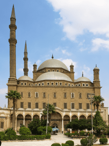 Exterior image of the Mosque of Mohammad Ali in Cairo Egypt. Two large minaret rise from the building on the left. A large white cloud is in the corner of the frame.